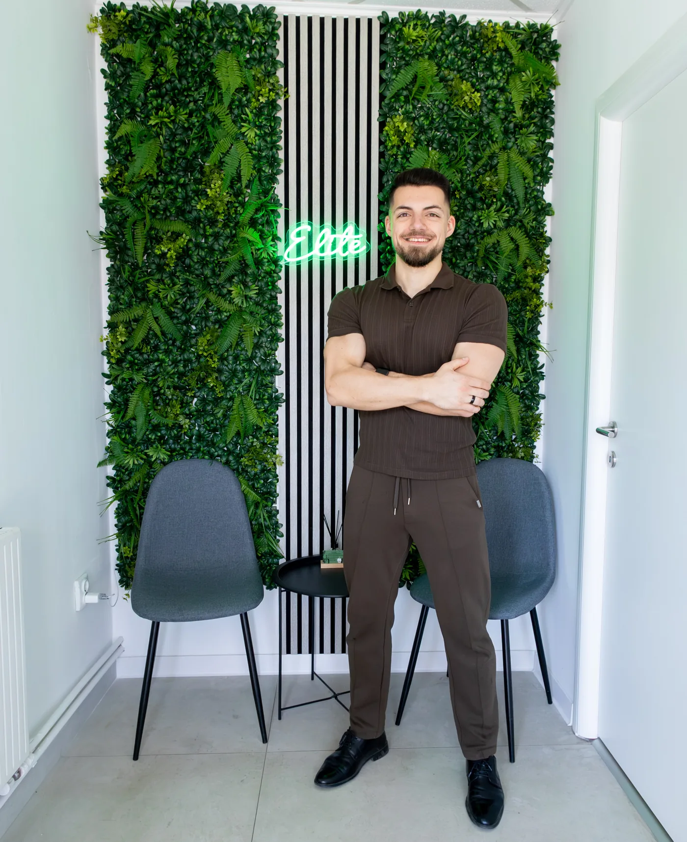 Balan Aurelian-Nicolae, kinetoterapeutul ELITE, zâmbind în fața peretelui de plante cu neonul Elite